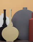 A set of minimalist ceramic vases in matte black, cream, white, gray, and terracotta red, arranged against a warm beige background.