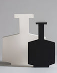 Two modern abstract ceramic vases with geometric shapes, one matte black in the foreground and one matte off-white/cream behind it, set against a neutral light gray studio background.