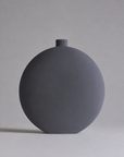 A matte dark gray ceramic vase with a small cylindrical neck and a rounded, disc-like body, photographed against a neutral light gray background.