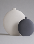 Two matte ceramic vases with rounded, disc-like shapes and short cylindrical necks, one in warm off-white and the other in dark charcoal gray, set against a neutral light gray background.
