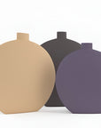 Three matte ceramic vases with a minimalist, rounded triangular shape and small cylindrical necks, shown in beige, dark gray, and purple against a clean white background.