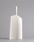 A minimalist white ceramic vase with a unique geometric, shield-like silhouette and a short cylindrical neck, standing on a neutral gray surface against a matching gray background.