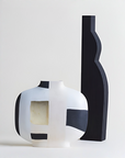 Two modern sculptural ceramic vases on a light gray background. One vase is white with black geometric blocks and a square inset showing a beige interior.
