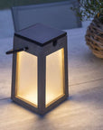 A LJ Solar Lantern Tecka Teck143 300L Duratek, with a solar module, is gracefully placed on a table beside a potted plant. The lantern features a Duratek® finish, enhancing its durability and longevity.