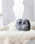 Two dark gray owl figurines with white speckled details, sitting on a fluffy cream sheepskin blanket draped over a white wooden chair.