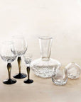 A set of clear glass diningware with textured hammered interiors and black bases, including a decanter, two wine glasses, a small tumbler, and a small vase, arranged on a light beige surface against a soft cream background.