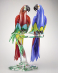 A colorful Murano glass sculpture by Zanetti of two parrots perched on a transparent glass branch with a round base, photographed against a light gray studio background, at Spacio Mumbai store in India.