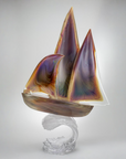 An calcedonio Murano glass sailboat sculpture with purple, gold, and brown marbled sails and hull, mounted on a clear wave-shaped glass base against a neutral gray studio background.