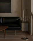 A tall silver floor candelabra with multiple candle holders and hanging crystal accents stands in a modern living room next to a black leather sofa and a wooden coffee table.