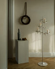 A tall silver metal candelabra with multiple candle holders and lit white candles stands in a softly lit room with wood flooring and neutral walls. A small pedestal with a vase and fruit is nearby, along with a dark woven wall hanging.