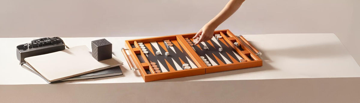 Hand reaching over a luxury backgammon board with leather handles, placed on a minimalist desk with books and decor.