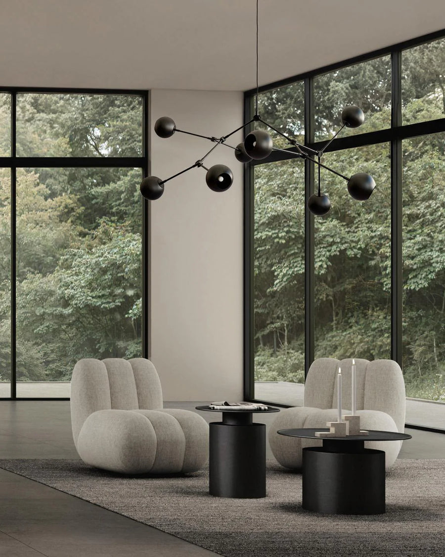 Modern living room with gray armchairs, black tables, and a decorative chandelier in front of large windows with a view of trees.