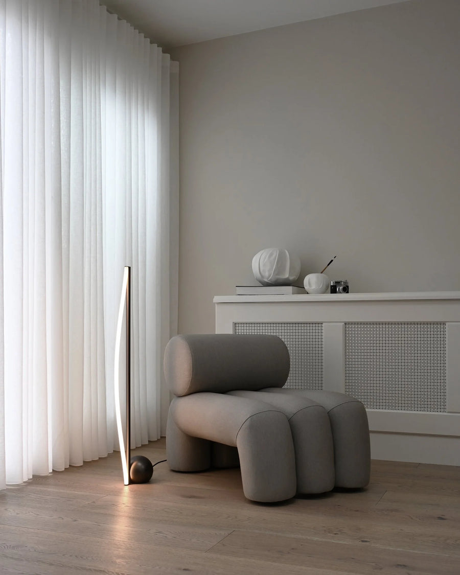 Modern gray armchair in a minimalistic room with light-colored curtains and wooden floor.