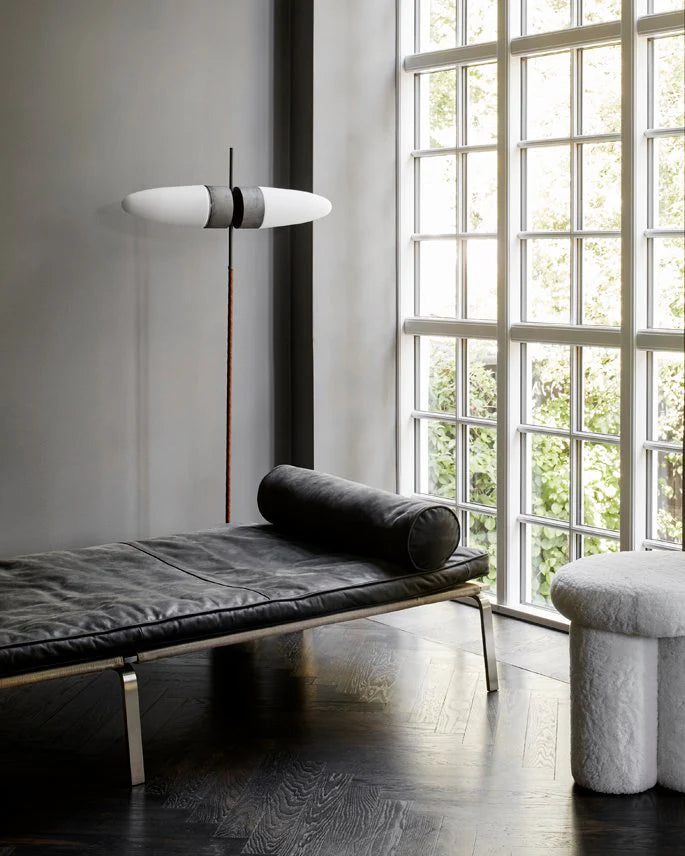 Modern interior with a black leather bench, gray ottoman, and floor lamp near a large window.