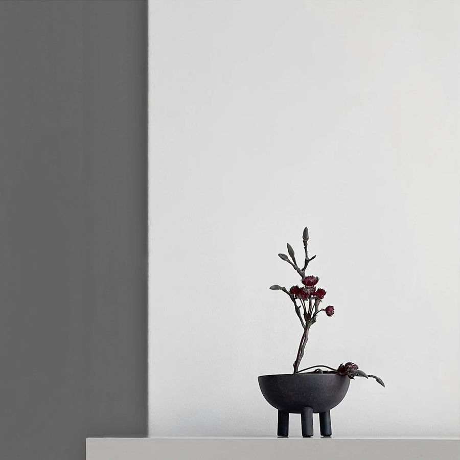 Coffee colour ceramic vase with branches and small flowers on a white surface against a gray wall.