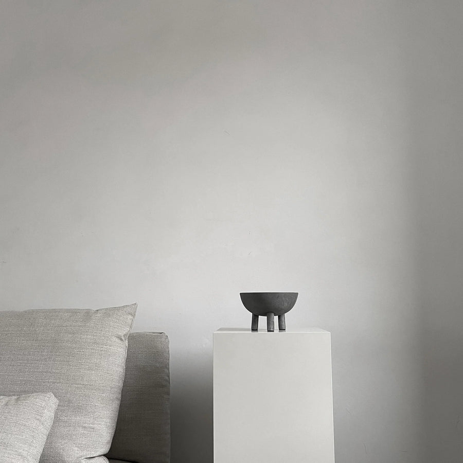 Dark grey colour bowl on a white pedestal against a plain gray wall with a gray sofa from 101 Copenhagen at Spacio retail stores in Mumbai & Kolkata. 