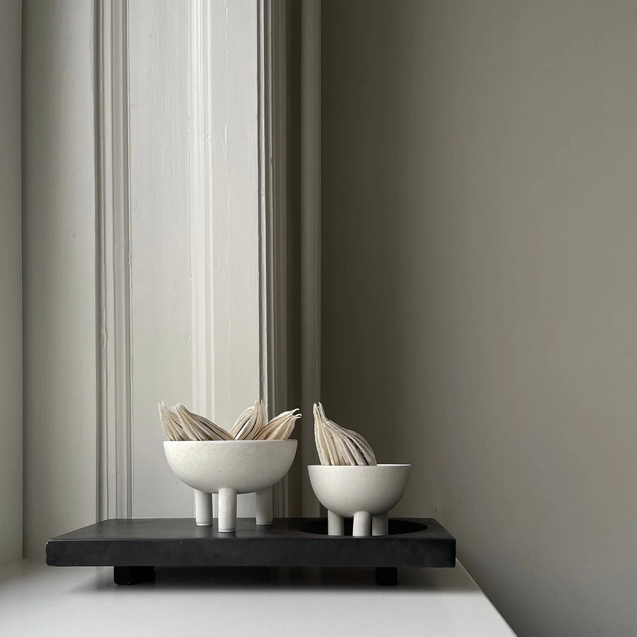 Two white ceramic bowls with plant stems on a black rectangular platform against a gray wall from 101 Copenhagen at Spacio retail stores in Mumbai & Kolkata. 