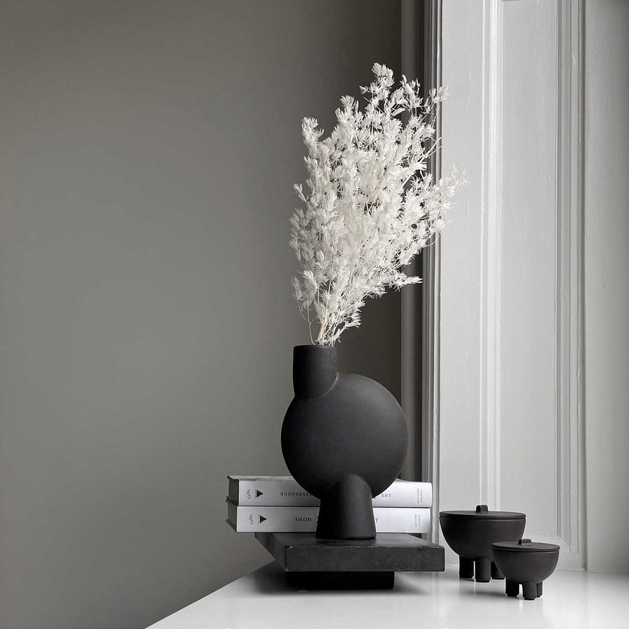 Black vase with dried white flowers on a gray wall background from 101 Copenhagen at Spacio retail stores in Mumbai & Kolkata. 