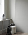 White stool with a gray towel against a light gray wall and a table lamp on windowsill. 