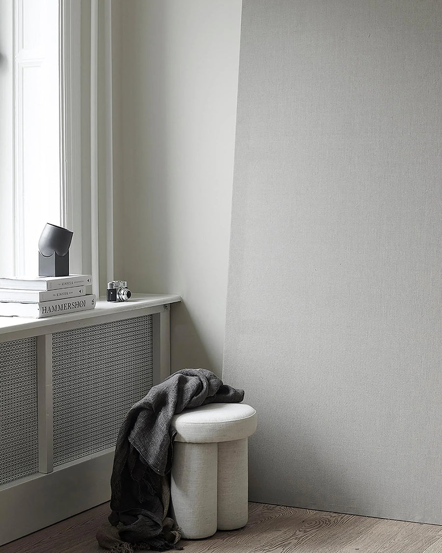 White stool with a gray towel against a light gray wall and a table lamp on windowsill. 