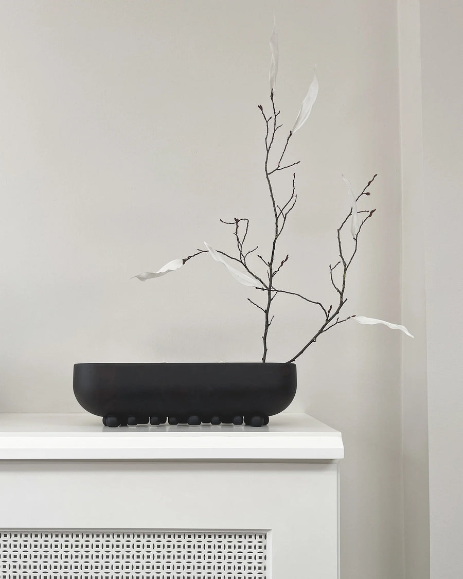 Black bowl with dried branches on a white surface against a light gray wall