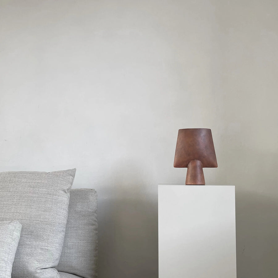 Ceramic vase from 101 Copenhagen on a white pedestal against a plain wall with a gray sofa.