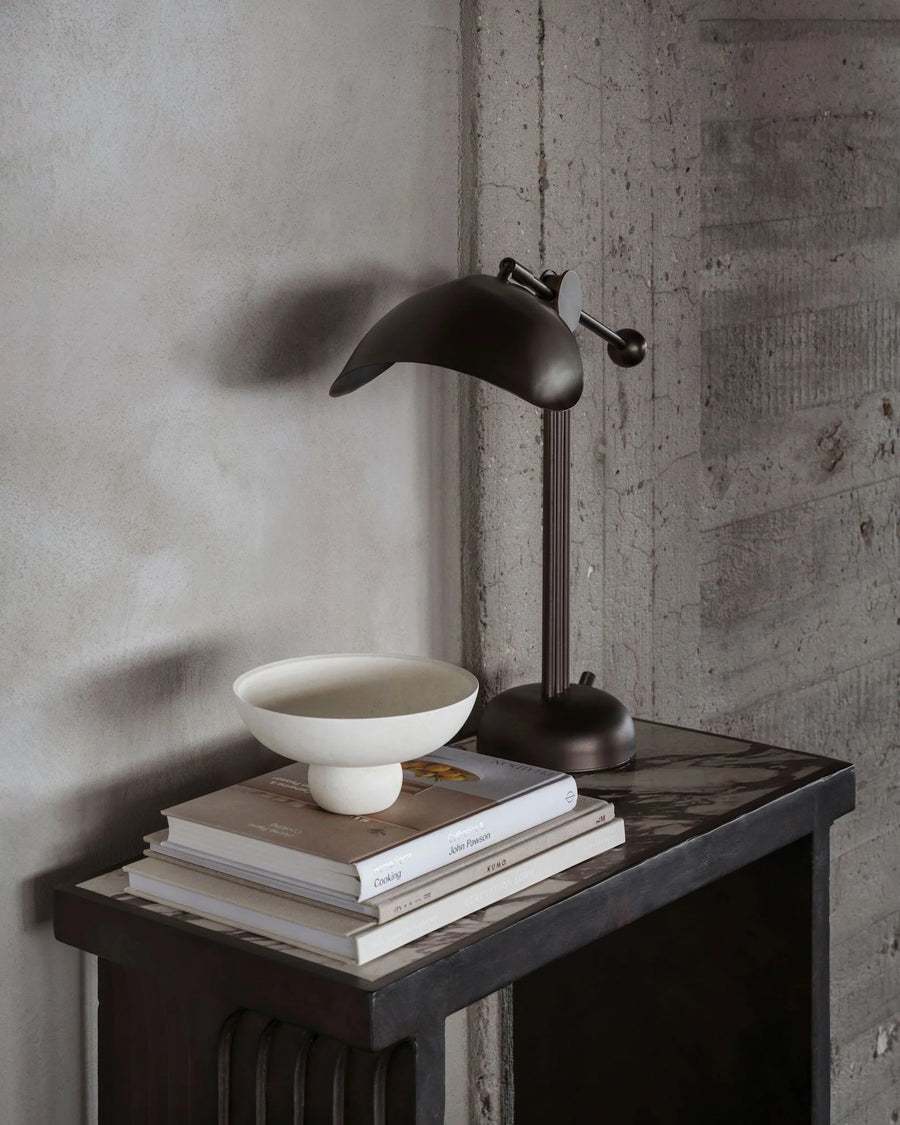 Dark metal desk lamp on a wooden surface with books and a bowl against a concrete wall.