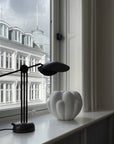 Modern black desk lamp on a windowsill with a white vase and building reflection.