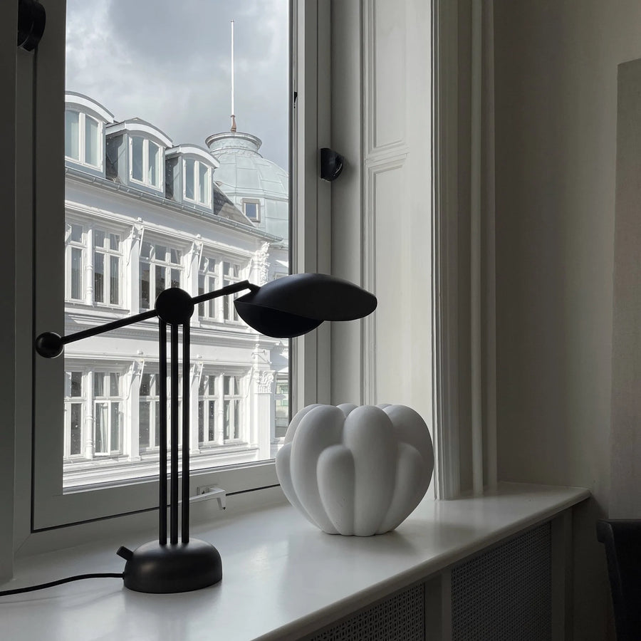 Modern black desk lamp on a windowsill with a white vase and building reflection.