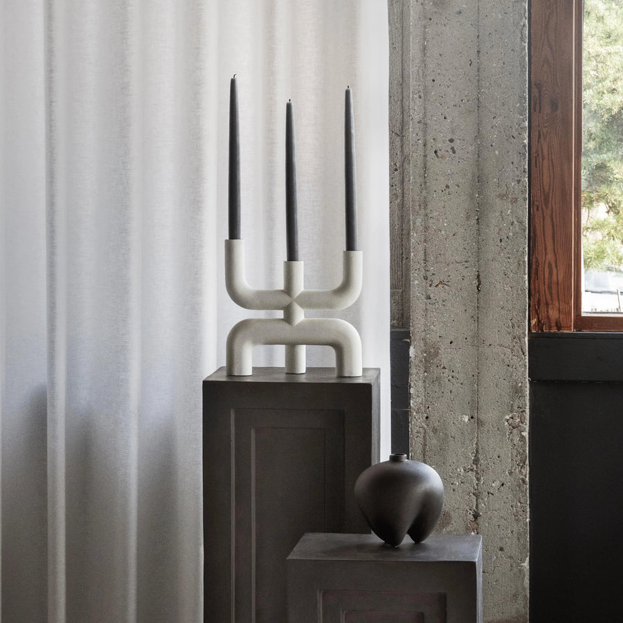 Modern candle holder with three black candles on a dark surface against a light curtain background from 101 Copenhagen at Spacio retail stores in Mumbai & Kolkata. 