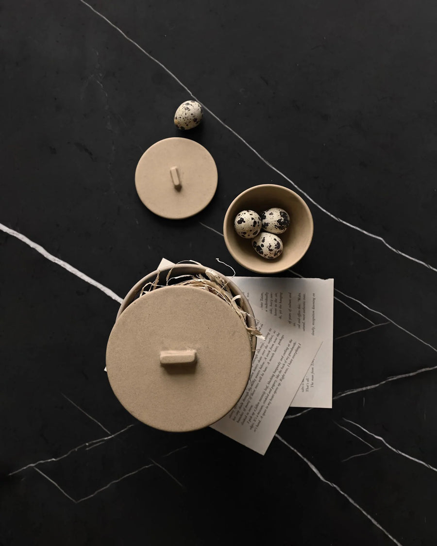 Small beige ceramic containers with lids on a dark surface from 101 Copenhagen at Spacio retail stores in Mumbai & Kolkata. 