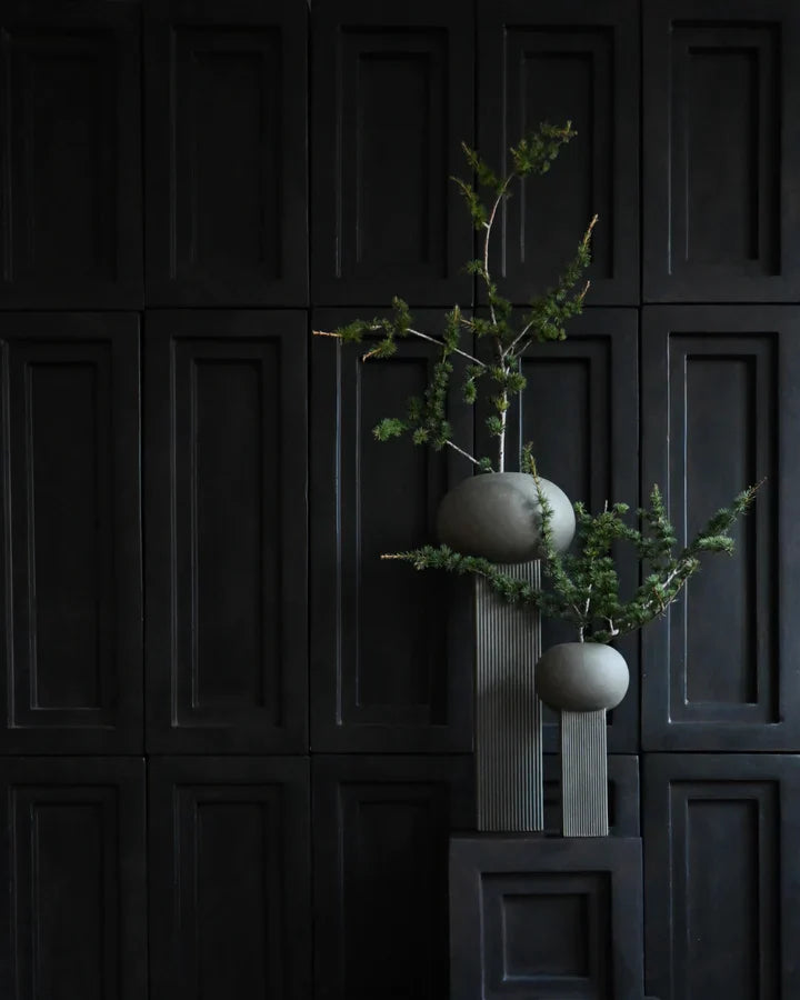 Two 101Cph Empire Mini Dark Grey 223036 ceramic vases with a plant on them in front of a black wall.