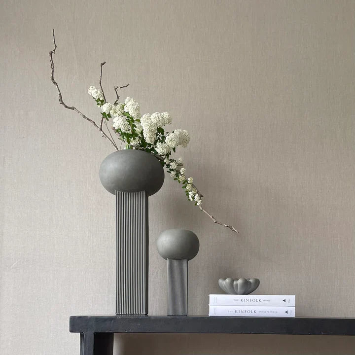 Two 101Cph Empire Mini Dark Grey 223036 ceramic vases by 101 Copenhagen, placed on a shelf alongside books and flowers.