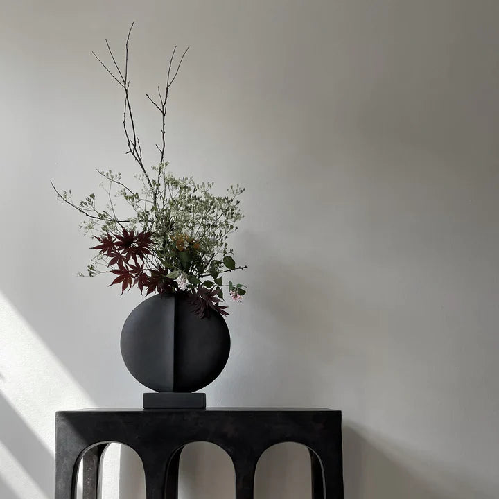 A quirky black vase, 101 Copenhagen Guggenheim Mini Coffee 111220, sits on a table in front of a white wall.