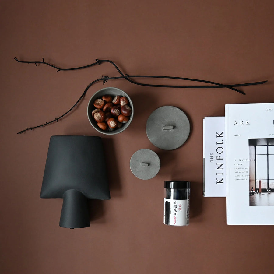 Decorative items including a black vase, gray containers, a bowl of nuts, and a magazine on a brown surface from 101 Copenhagen at Spacio retail stores in Mumbai & Kolkata. 