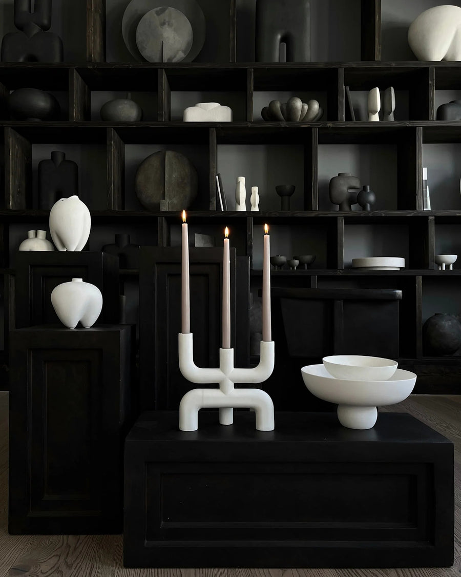Modern candle holder with white base and black candles on a dark surface with shelves in the background from 101 Copenhagen at Spacio retail stores in Mumbai & Kolkata. 