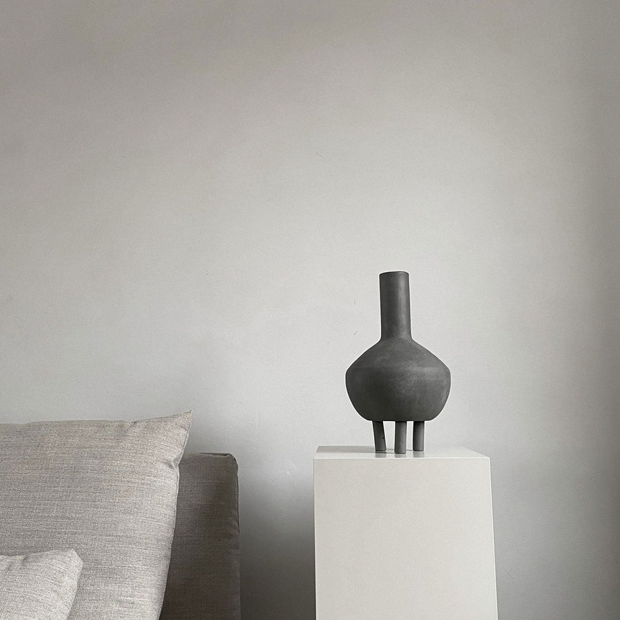 A 101Cph Duck Fat Dark Grey 111281 vase sits on a pedestal in front of a couch.