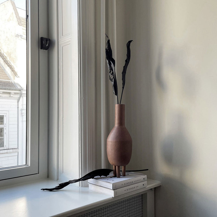 A 101Cph Duck Vase Slim Terracotta 213044, made by 101 Copenhagen, sits on a window sill next to a book.