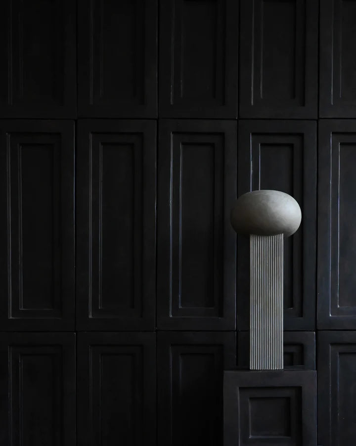 An architectural reference of a ceramic 101Cph Empire Big Dark Grey 223034 vase, resembling a concrete ball, elegantly sits on a pedestal in front of a black wall.