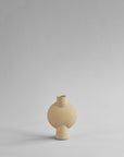 A beige vase with a sand finish sitting on a gray surface. This Scandinavian brand, 101 Copenhagen, offers the stylish 101Cph Sphere Bubl Mini Sand 203008 as an elegant home decor piece.