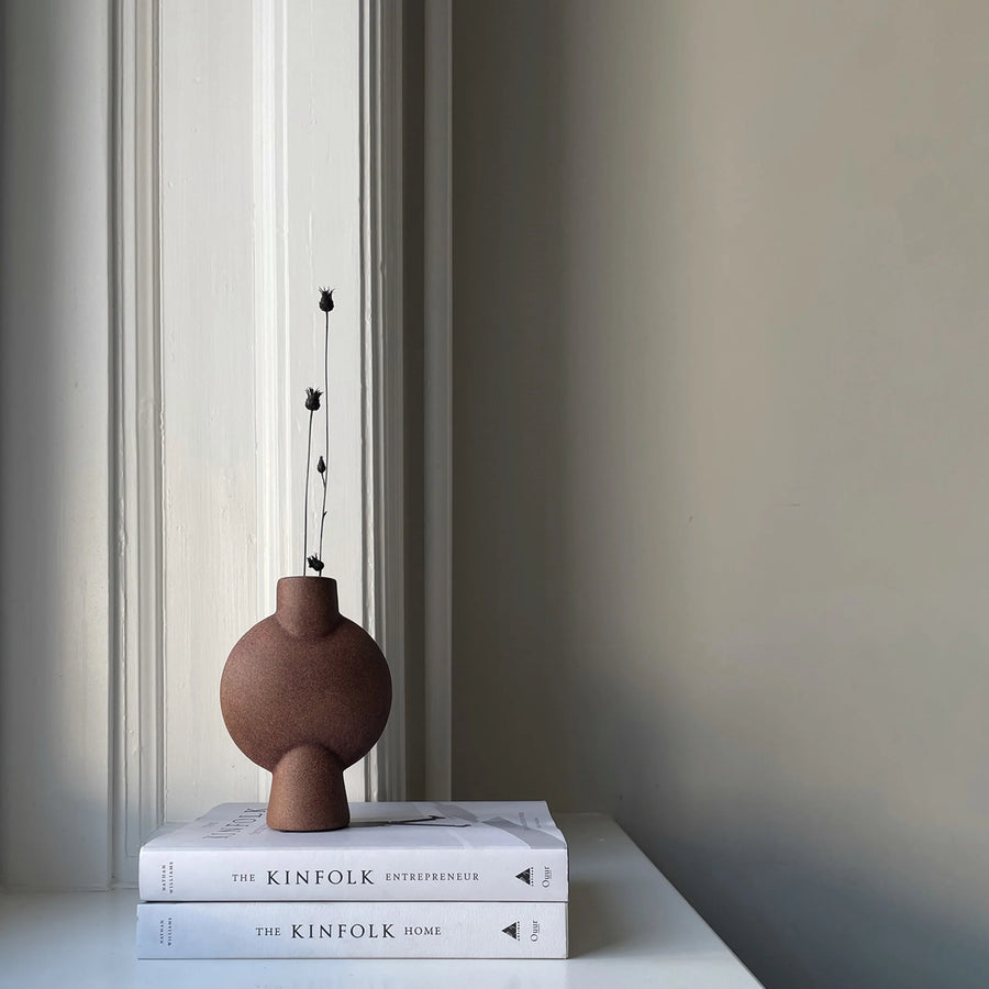 A 101Cph Sphere Bubl Mini Terracotta 203010 vase sits on top of a bookshelf next to a window, manufactured by 101 Copenhagen.
