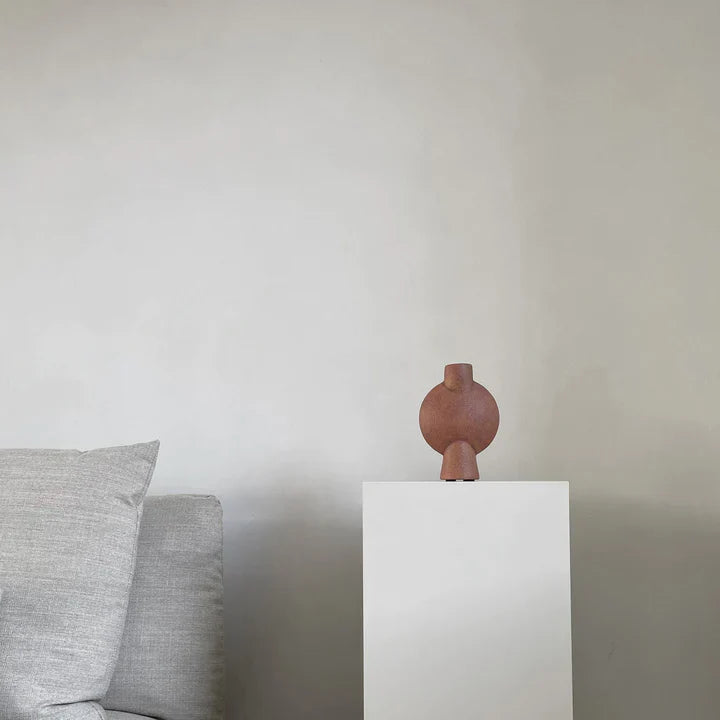 A brown 101Cph Sphere Bubl Mini Terracotta 203010 vase from the Scandinavian brand 101 Copenhagen sits on a white pedestal in a living room.