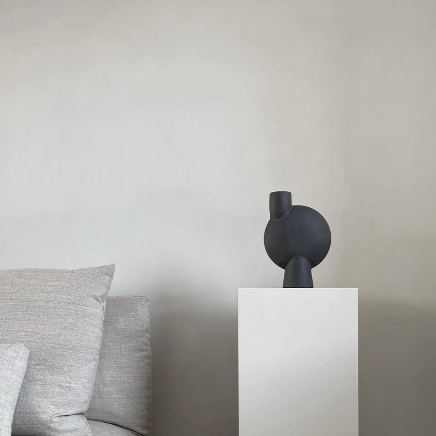 Black abstract sculpture on a white pedestal against a neutral wall with gray cushions from 101 Copenhagen at Spacio retail stores in Mumbai & Kolkata. 
