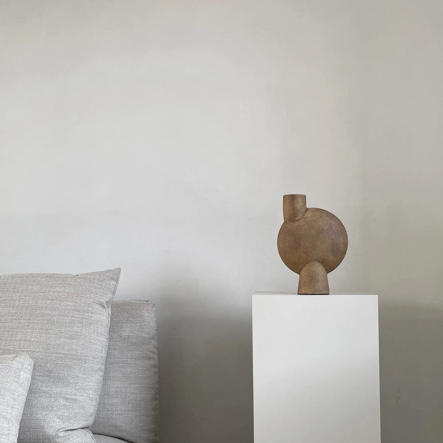 Decorative vase on a white pedestal against a plain wall with gray cushions from 101 Copenhagen at Spacio retail stores in Mumbai & Kolkata. 