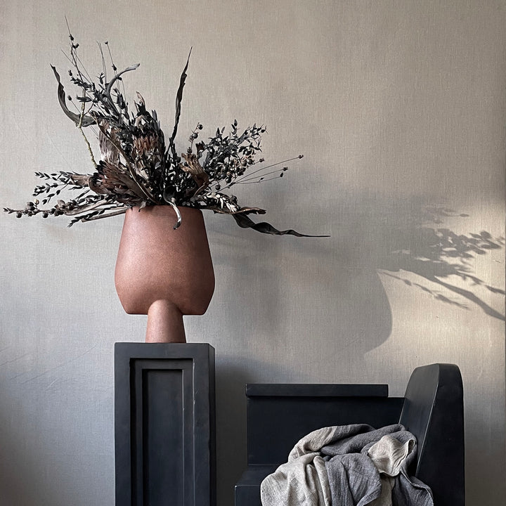 A 101 Copenhagen brand vase, the 101Cph Sphere Vase Square Big Terracotta 212008, with a Terracotta finish, placed on a table next to a chair.