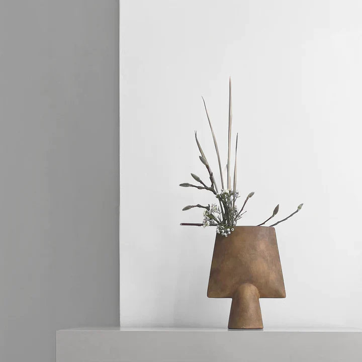 A 101Cph Sphere Vase Square Mini Ocher 111181 by 101 Copenhagen with an ochre finish sitting on top of a white wall.