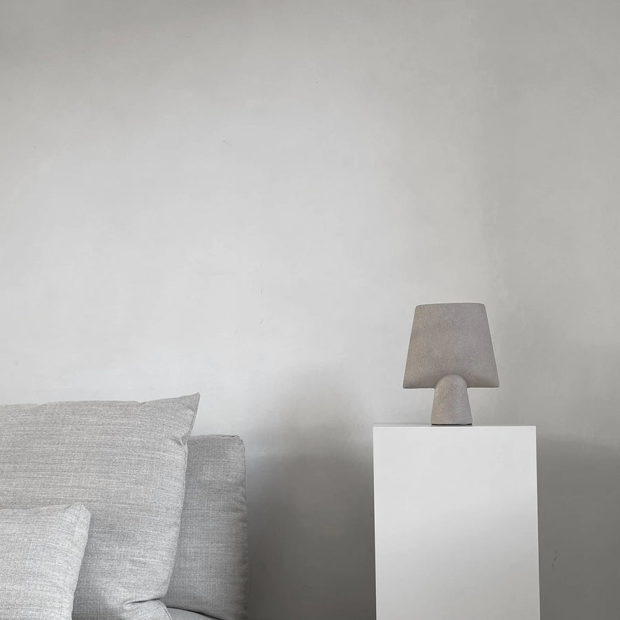 Gray ceramic vase on a white pedestal against a plain wall with gray cushions from 101 Copenhagen at Spacio retail stores in Mumbai & Kolkata. 