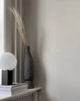 A 101 Copenhagen table lamp with a dark grey finish sits on a window sill next to a book, creating a perfect blend of Scandinavian living style.