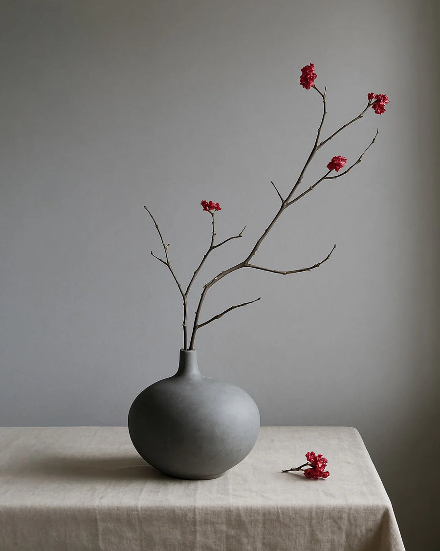 Gray vase with branches and red flowers on a beige tablecloth against a gray background from 101 Copenhagen at Spacio retail stores in Mumbai & Kolkata. 