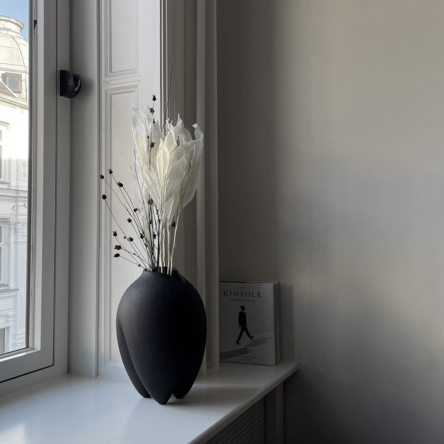 Black vase with dried plants on a windowsill next to a book  from 101 Copenhagen at Spacio retail stores in Mumbai & Kolkata. 